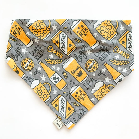 Image of Luca and B's elastic dog bandana in the "Beers" pattern; a dark grey background with golden beer bottles, glasses, cans, and mugs, bottle openers, wheat grass, splashes, white stars, and the words "lager, craft beer, IPA, porter, beer, ale" in a black, handwritten script.