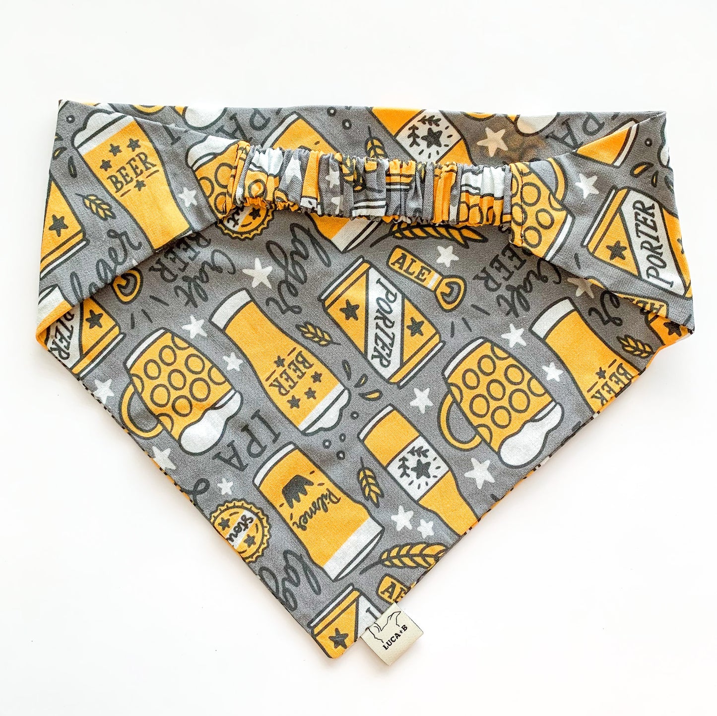 Image of the back and scrunchie piece of Luca and B's elastic dog bandana in the "Beers" pattern; a dark grey background with golden beer bottles, glasses, cans, and mugs, bottle openers, wheat grass, splashes, white stars, and the words "lager, craft beer, IPA, porter, beer, ale" in a black, handwritten script.